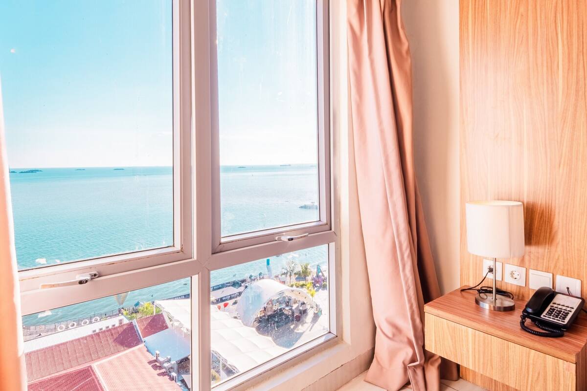Een hotelkamer met een groot raam met uitzicht op een strand en de zee.