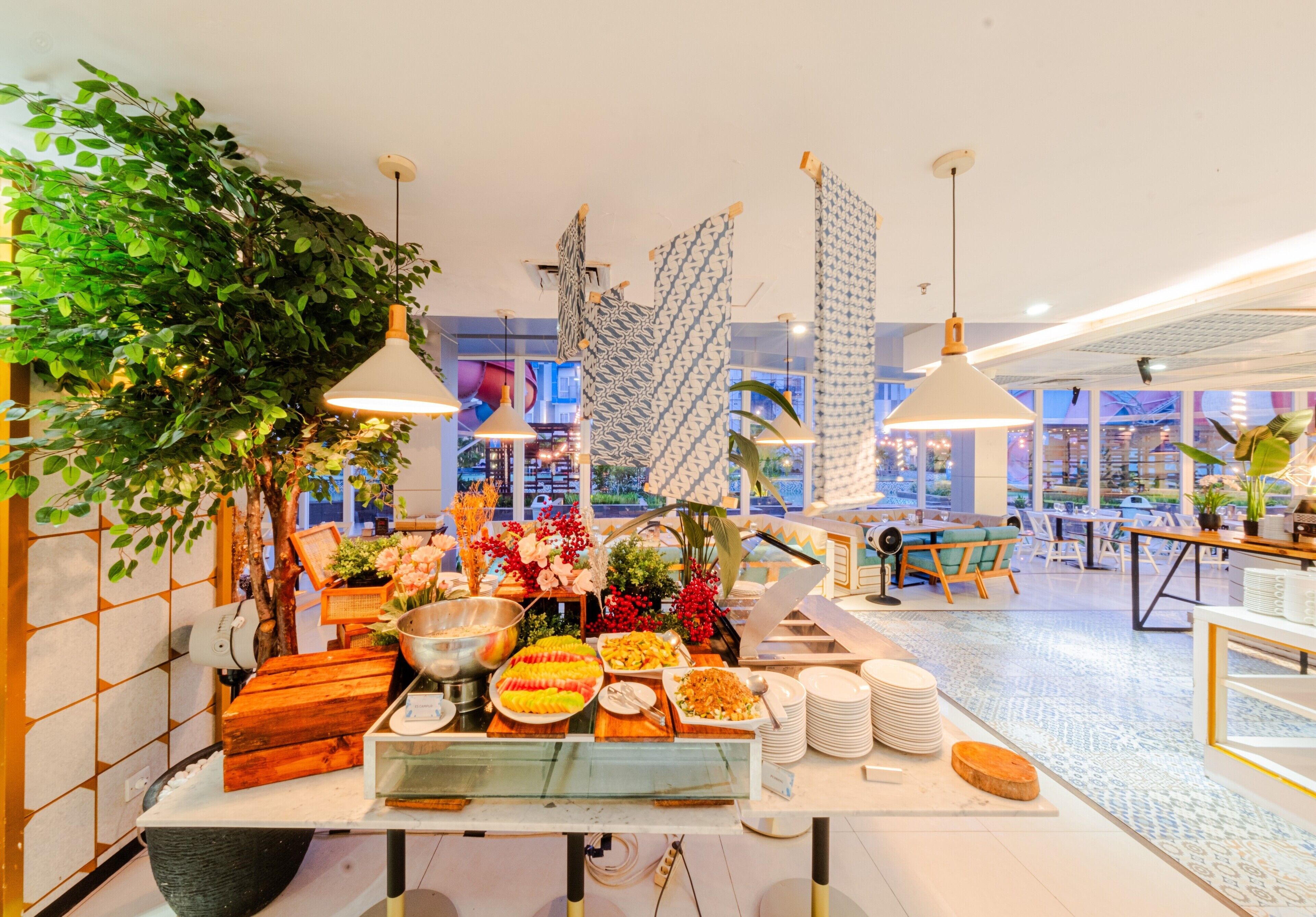 Mesa de buffet con diferentes opciones de comida, una planta grande con follaje y un interior moderno con lámparas colgantes y vistas a la ciudad.