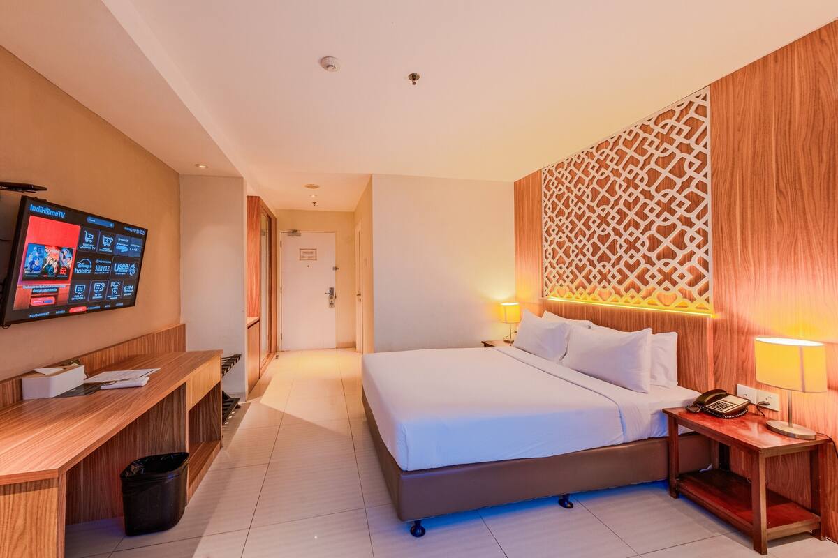 Een hotelkamer met een bed, nachtkastje, lamp en een aan de muur bevestigde televisie.