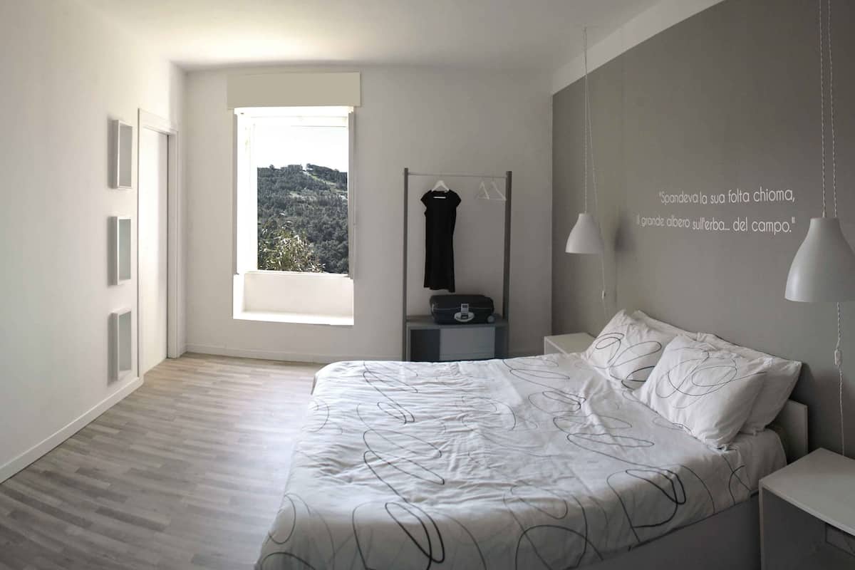 Un dormitorio con un ventanal grande, una cama con colcha blanca, dos mesitas de noche, una lámpara colgante y un espejo montado en la pared.