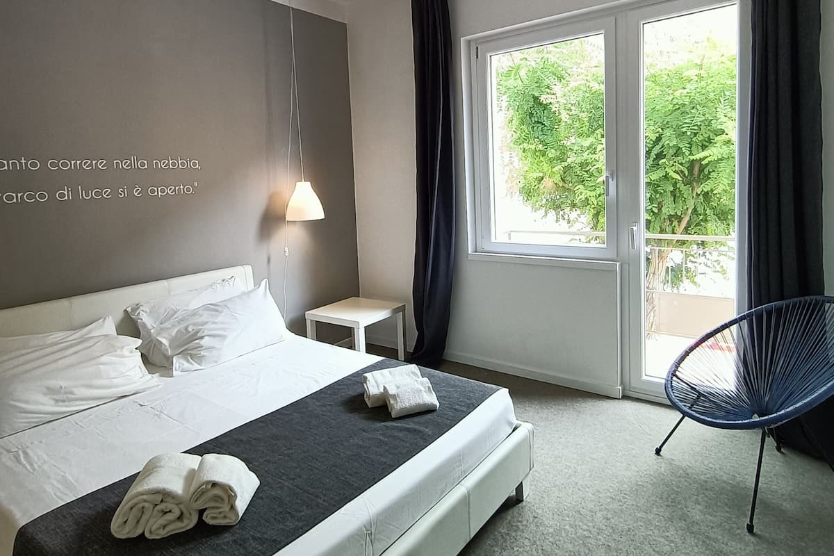 Habitación de hotel con cama, una mesita, una silla y un ventanal con cortinas.