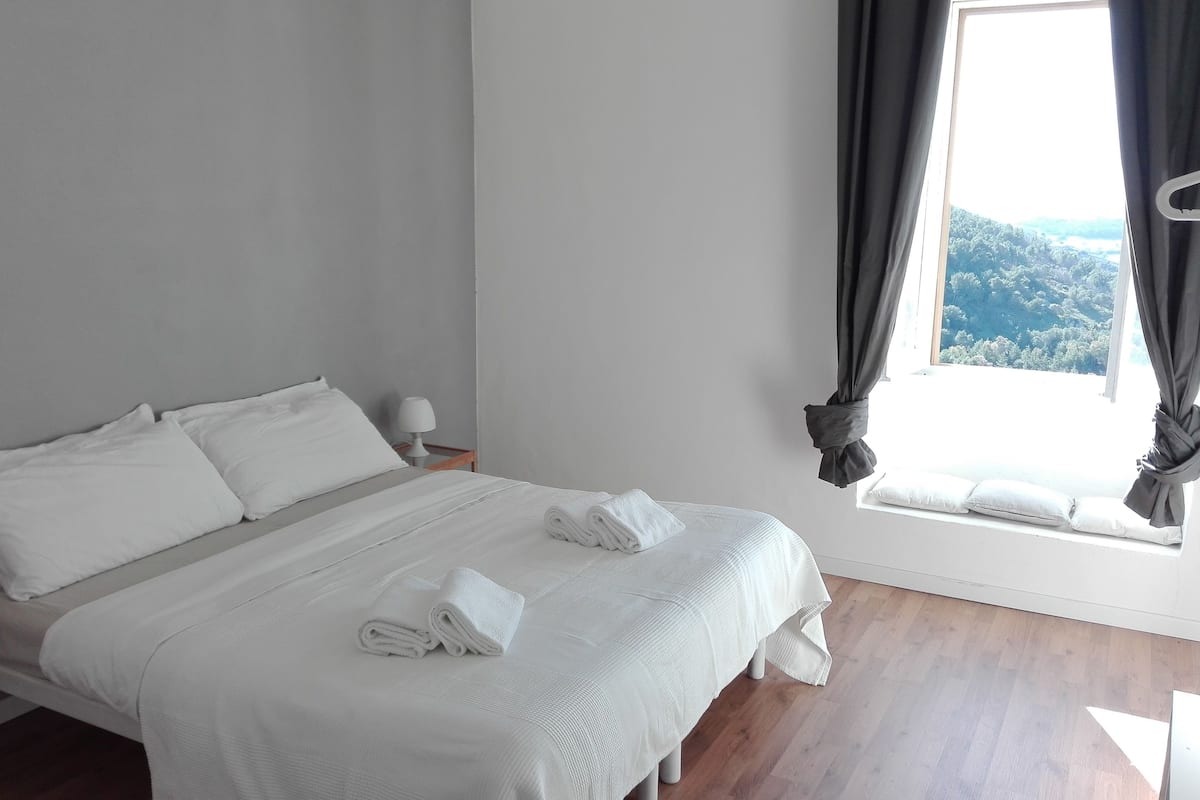 Una cama bien hecha con sábanas y toallas blancas, una mesita de noche con lámpara y una ventana con cortinas.
