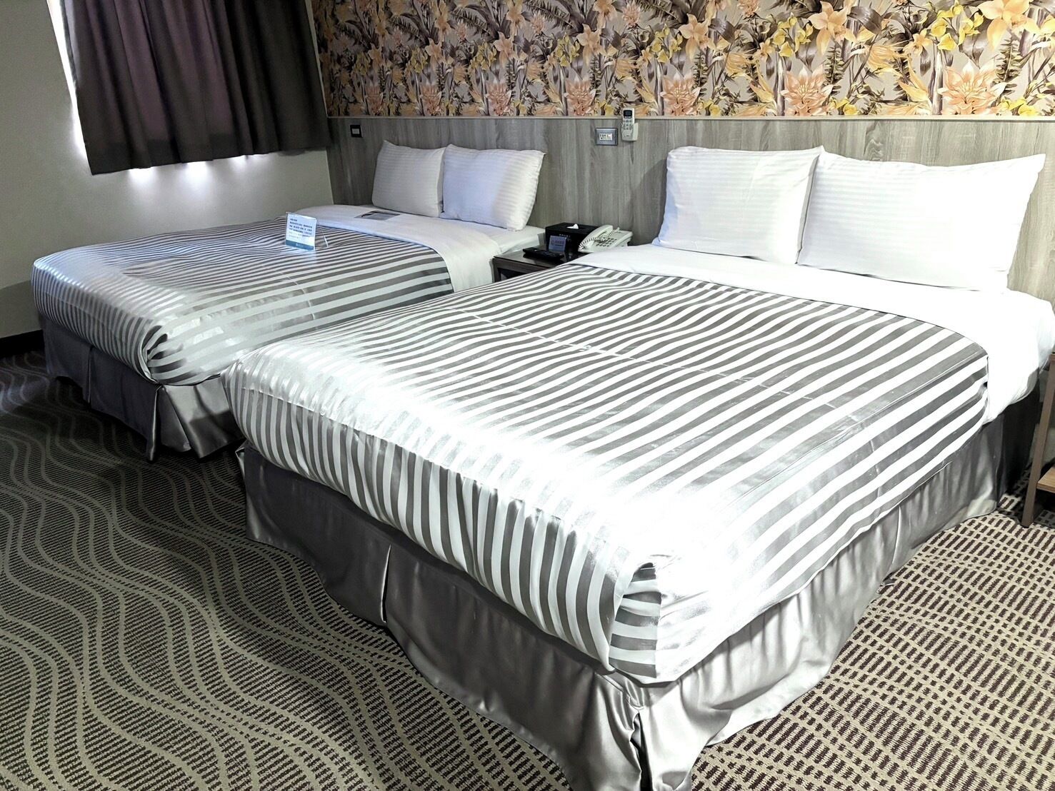 A hotel room with two beds, a patterned carpet, and a floral wallpaper.
