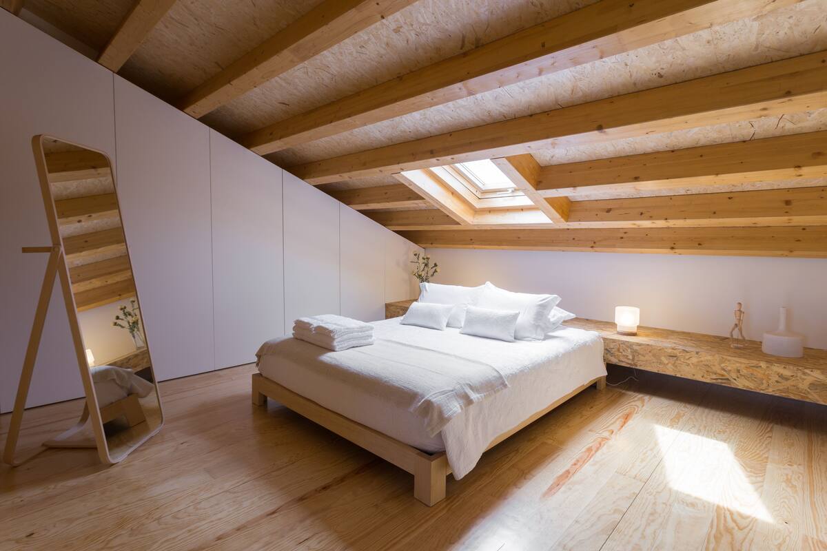 Quarto com teto de madeira, claraboia, cama com lençóis brancos e mesa de cabeceira com luminária.