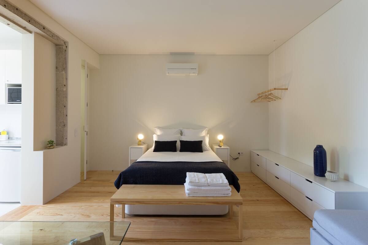 Quarto minimalista com cama, mesas de cabeceira, abajur, um banco de madeira e um cabideiro.