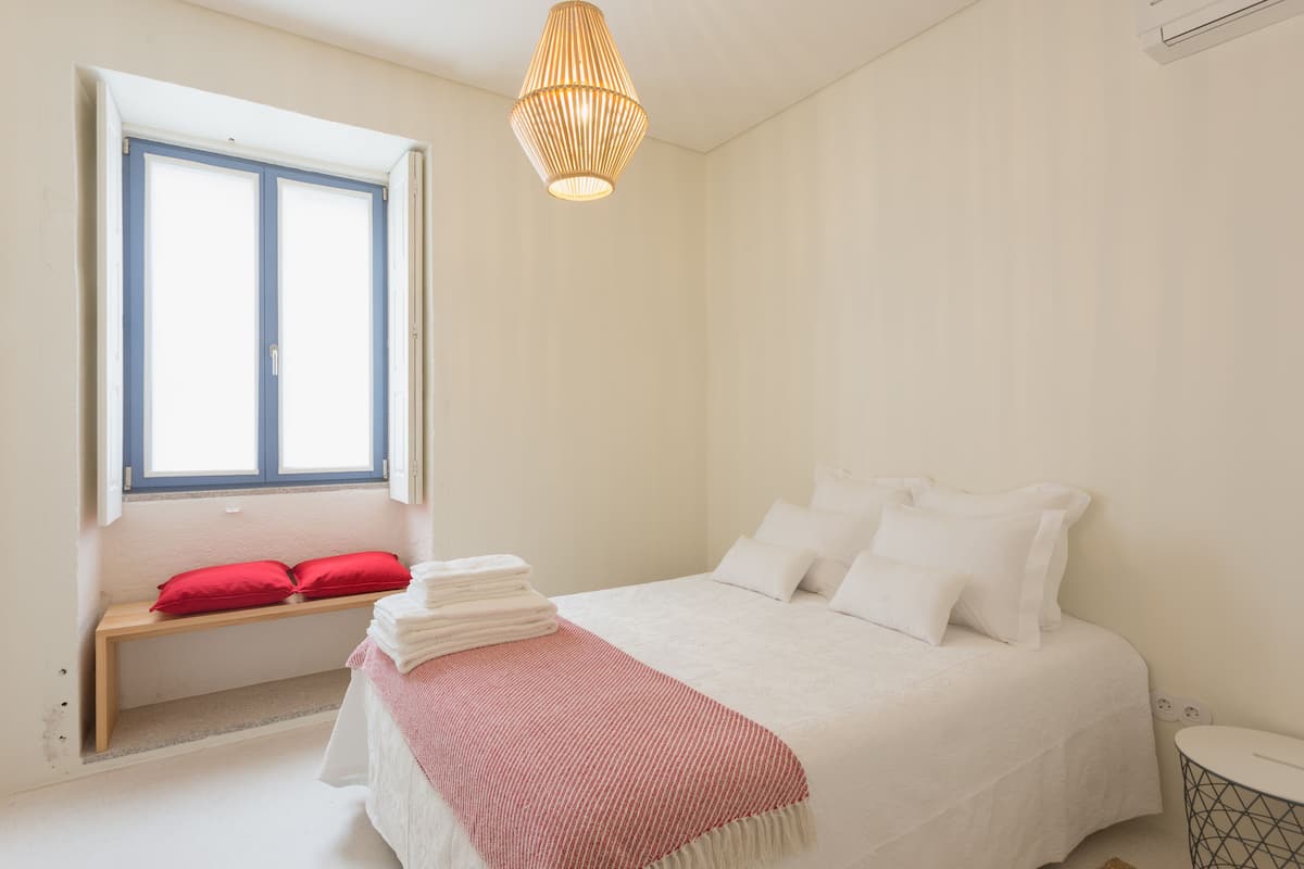 Um quarto com cama, janela, um banco e mesa de cabeceira.