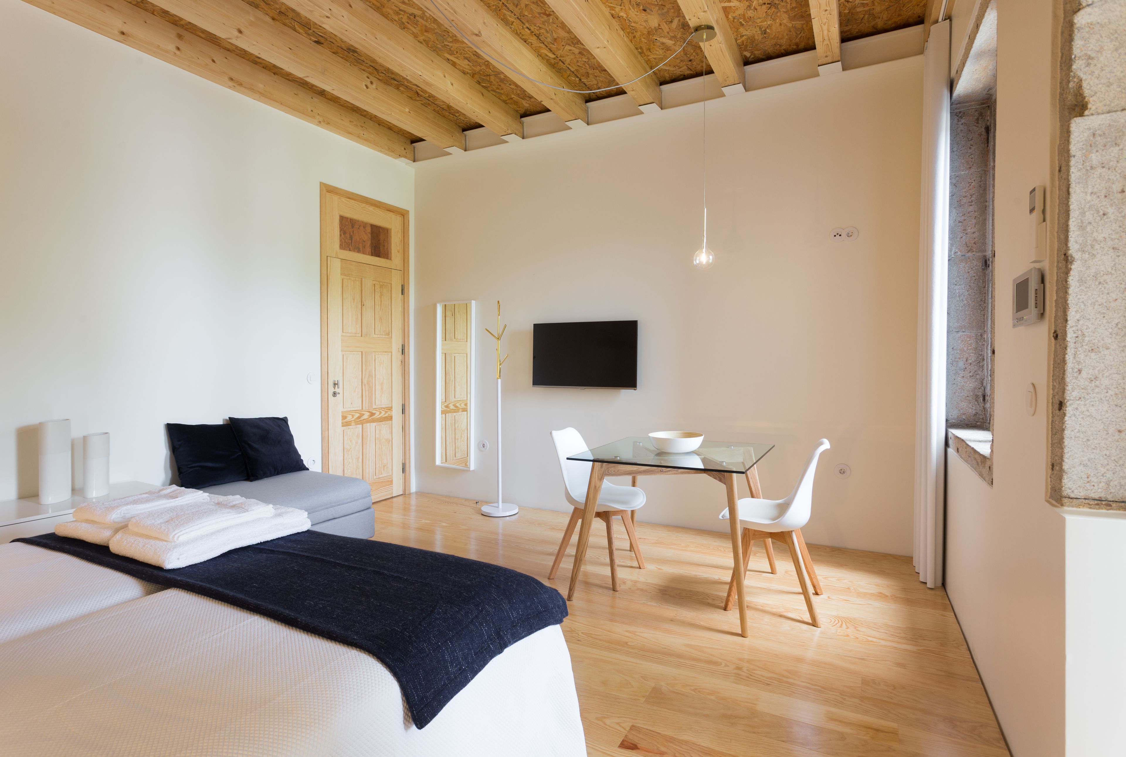 Een moderne slaapkamer met een bed, nachtkastjes, een flatscreen televisie, een glazen eettafel met stoelen en een houten plafond.