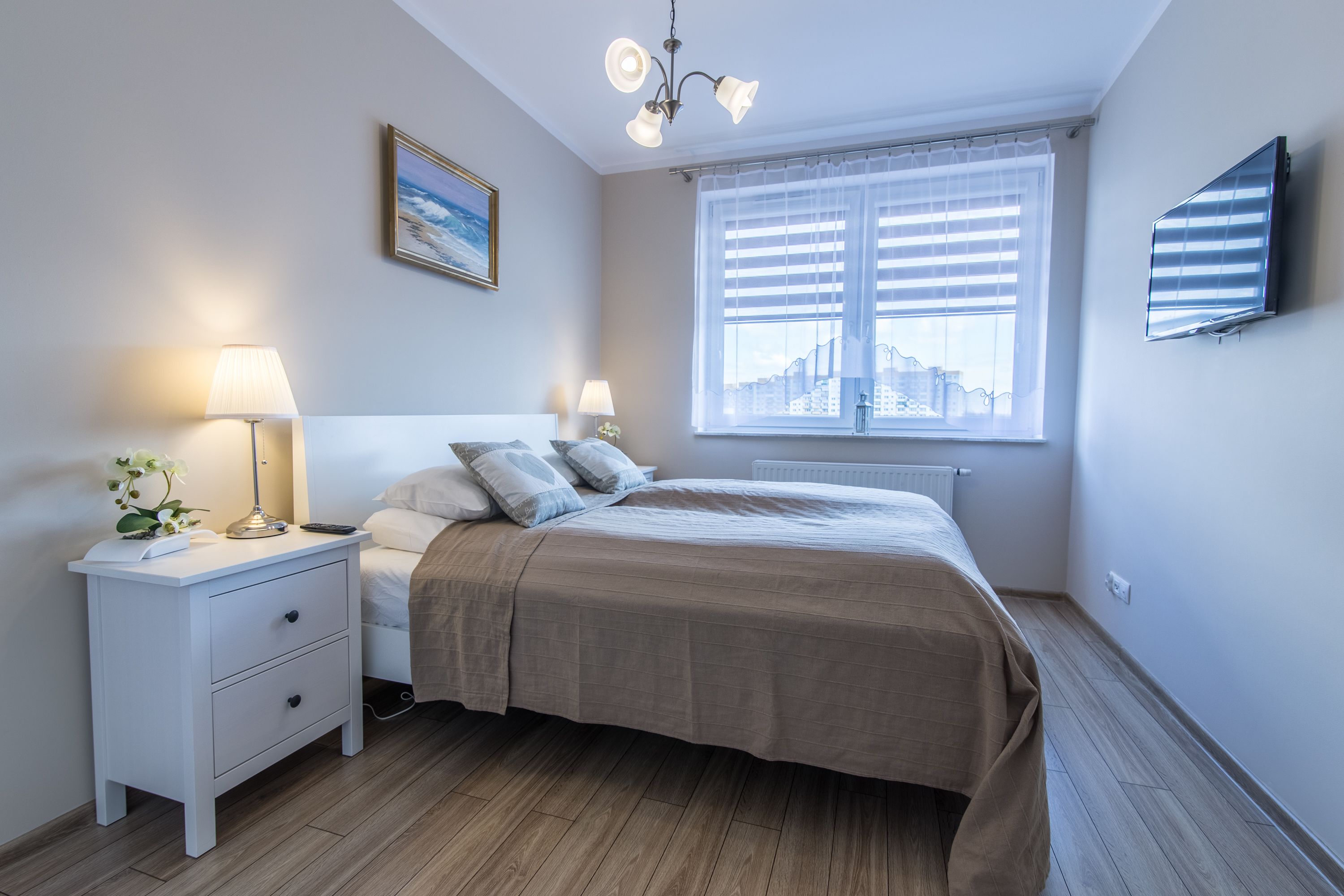 Quarto com cama, abajures, mesa de cabeceira, quadro emoldurado, janela com persianas e TV fixada na parede.
