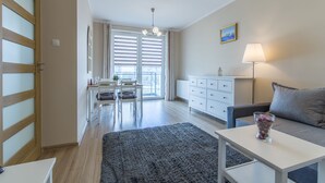 A modern living room with a sofa, a coffee table, a lamp, and a view of the dining area through a large window. - Rent a Flat Apartments - Jana Pawla II (Gdansk)