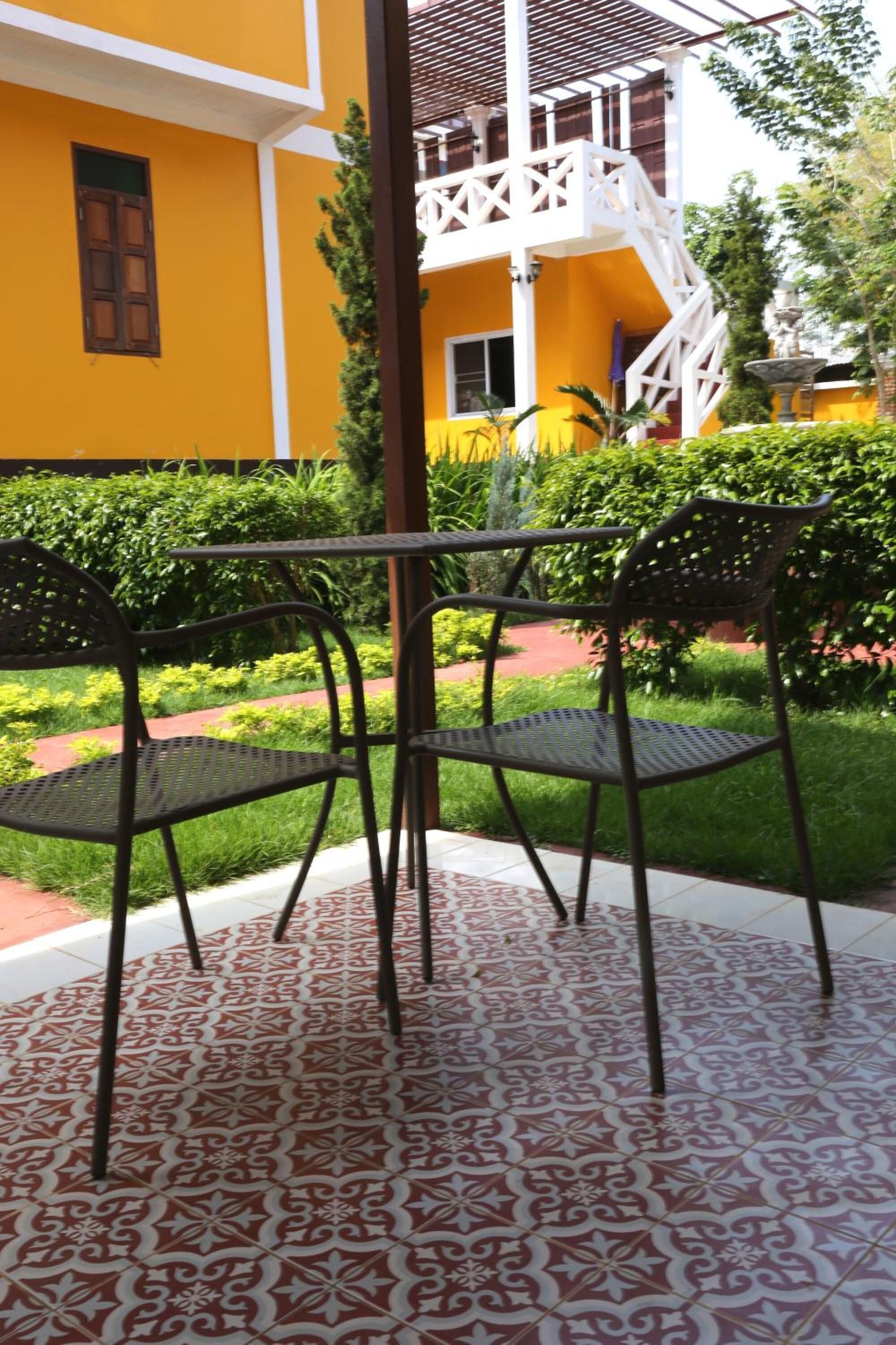Eine Terrasse mit Metallstühlen und einem gemusterten Boden, umgeben von einem gelben Gebäude und viel Grün.