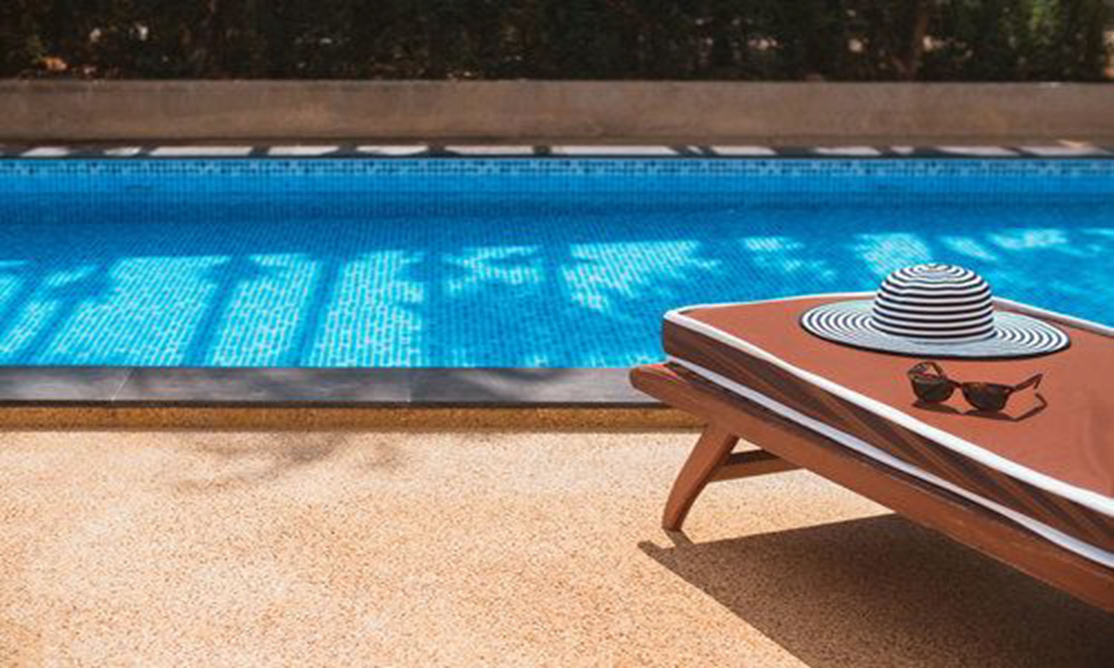 Una silla de descanso junto a la piscina, con un sombrero a rayas y gafas de sol, una piscina con azulejos y un borde de hormigón.