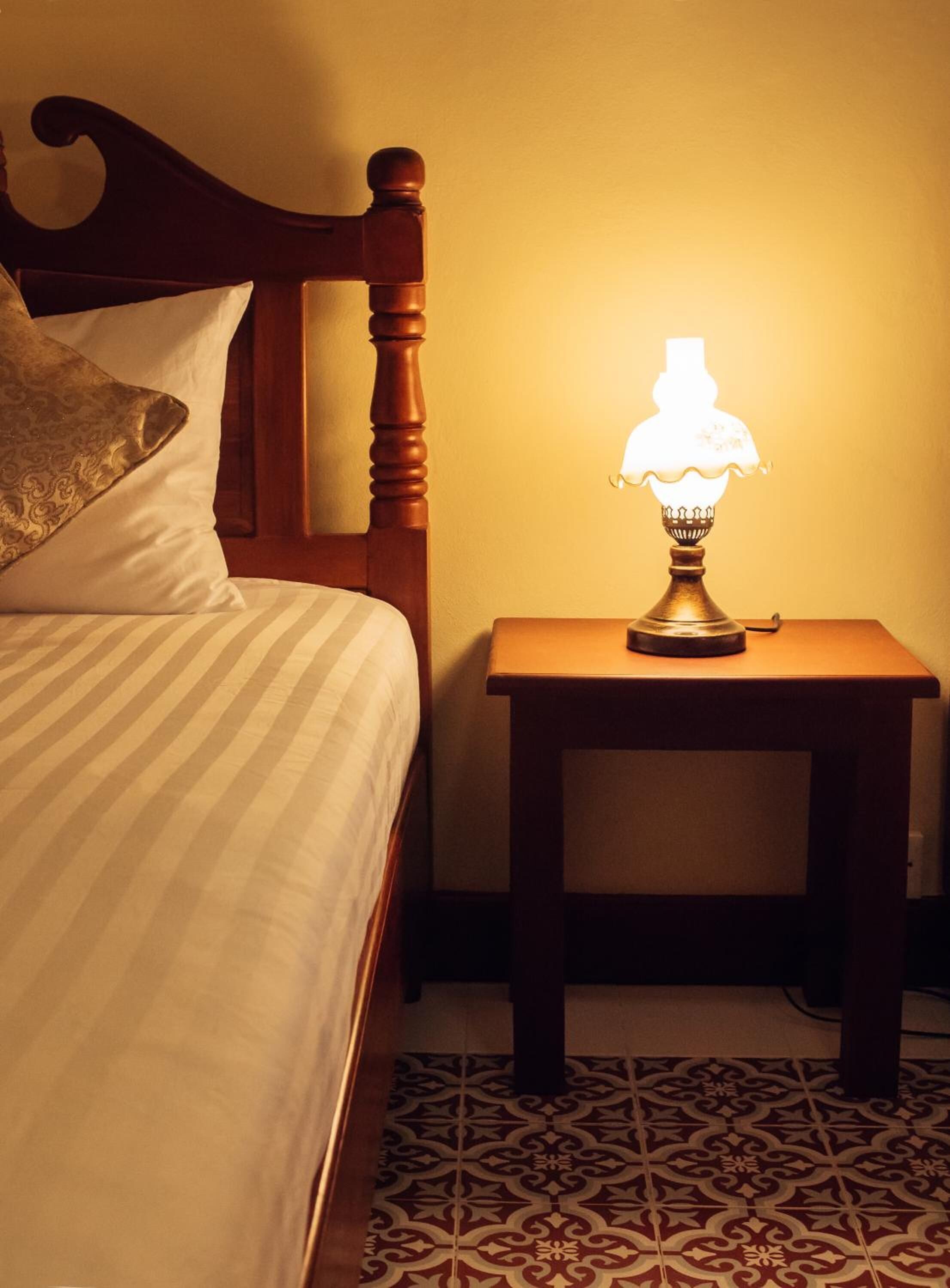 A neatly made bed with striped bedding and a decorative pillow, a wooden nightstand with a table lamp, and a patterned floor.
