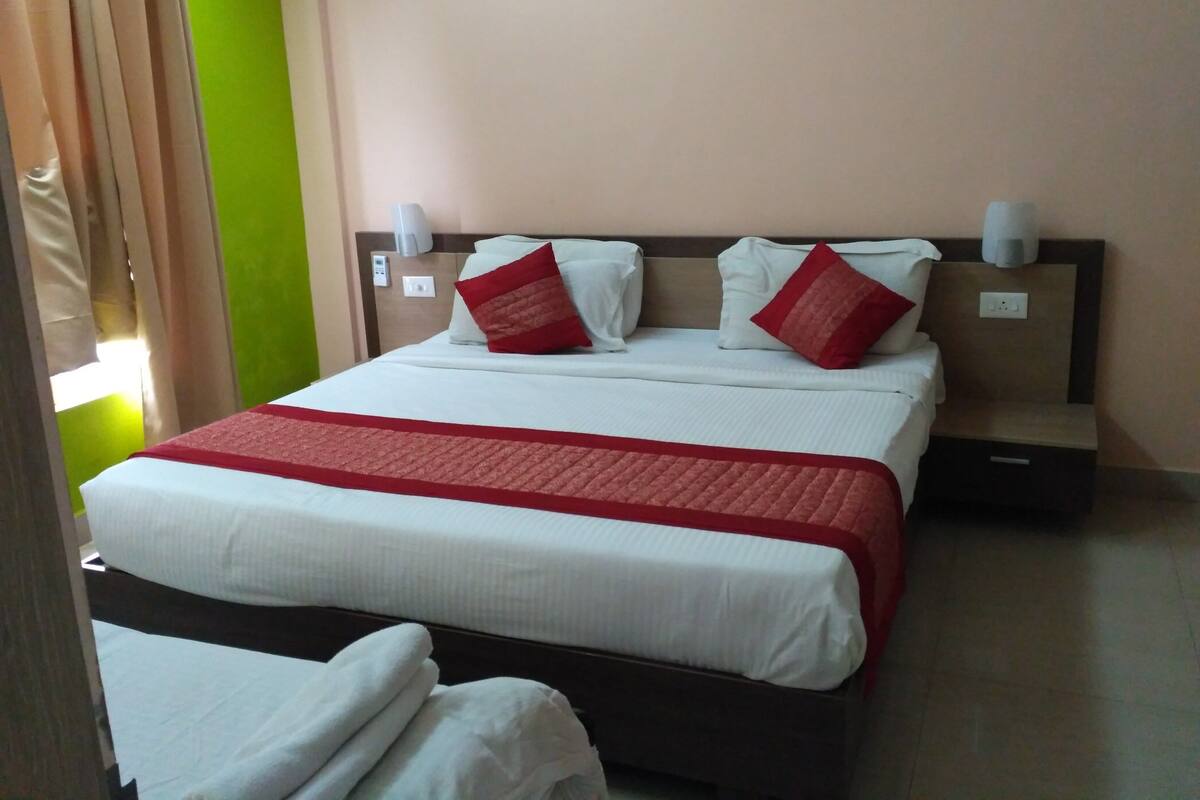 A hotel room with a large bed, white bedding, red accent pillows, a red throw blanket, a nightstand, and a wall-mounted lamp.