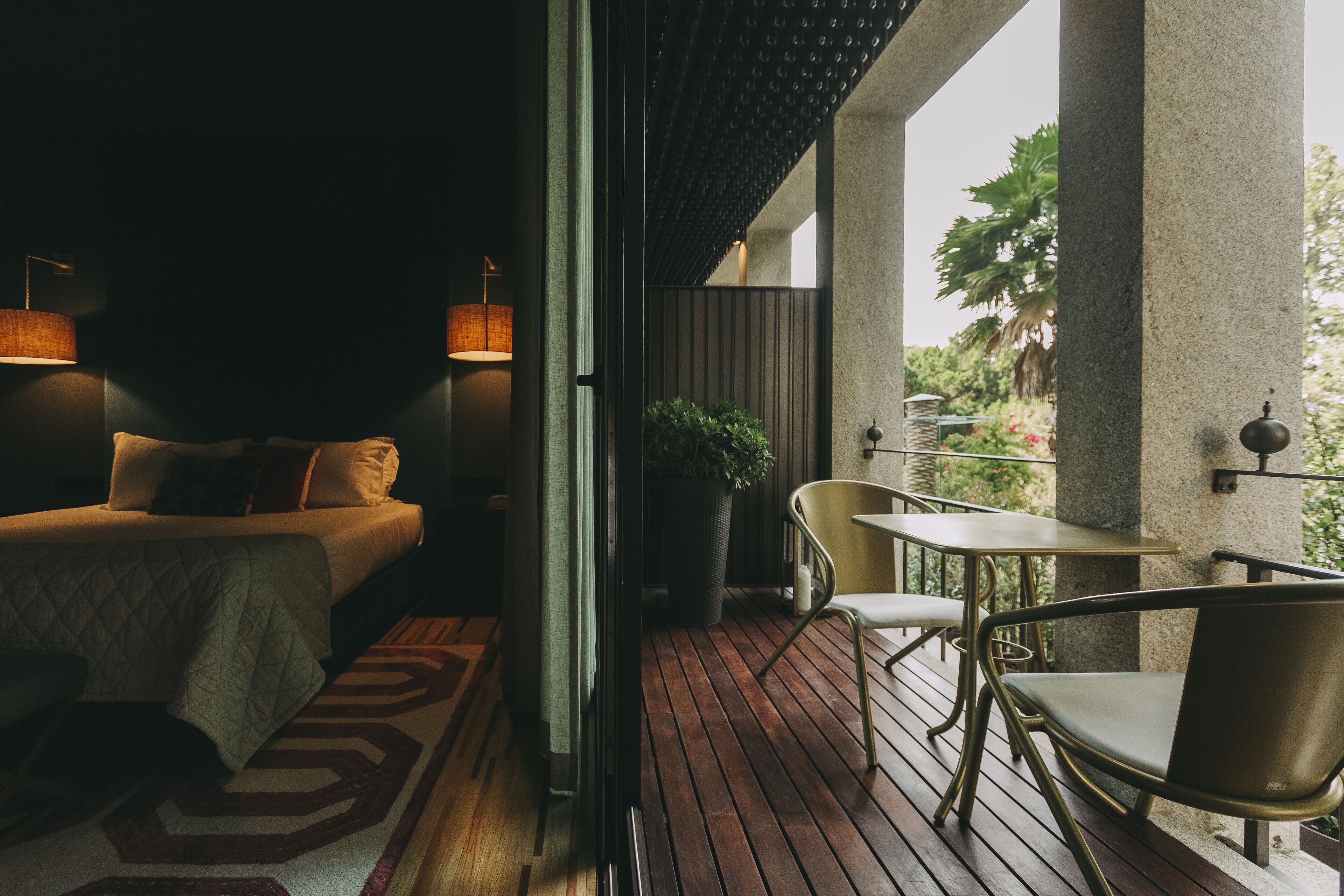 A hotel room with a bed, bedside lamps, a small table, and a chair. There is a potted plant and a view of greenery outside.