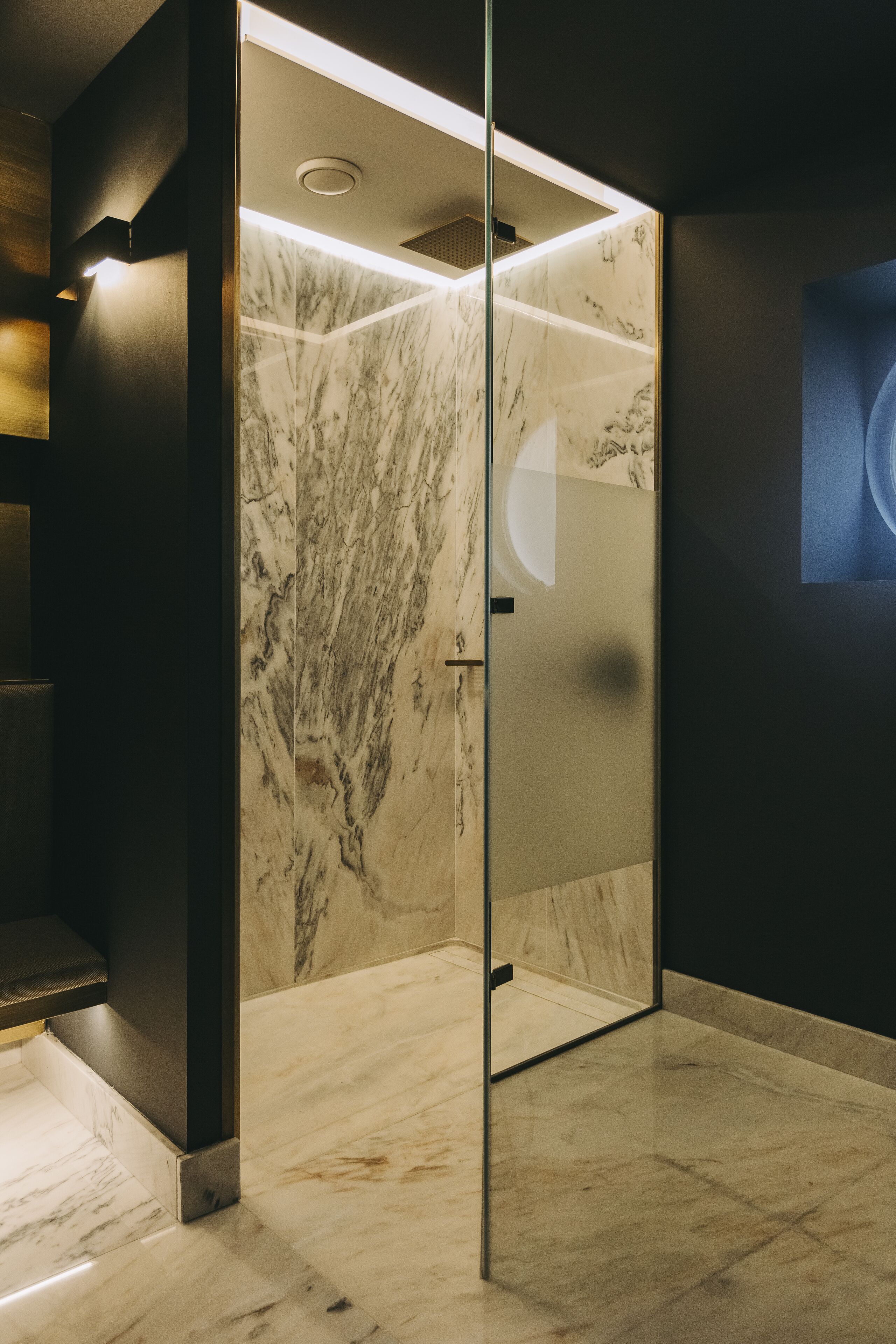 A modern elevator with marble walls and a glass door, featuring a ceiling light and a small vent.