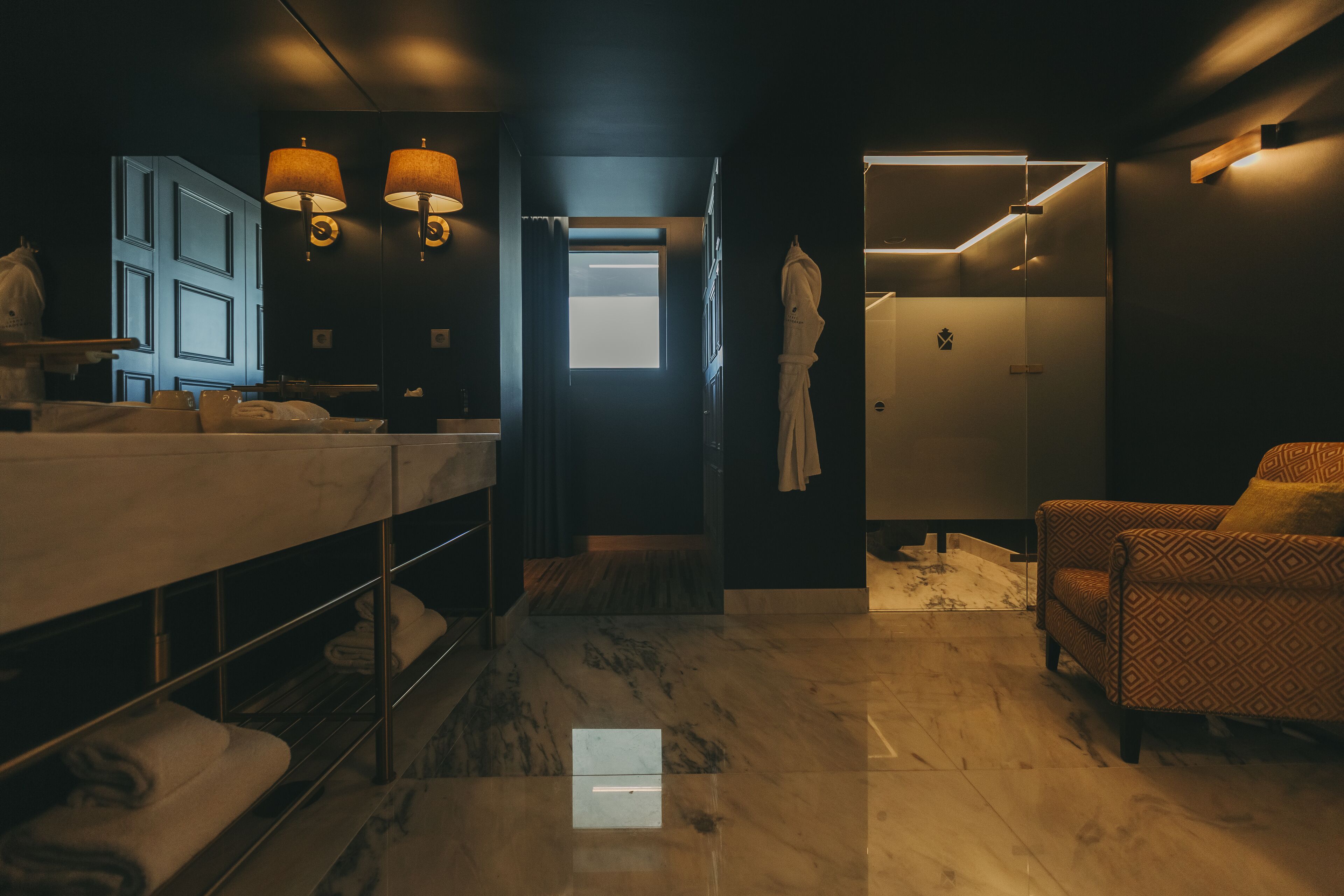 A modern bathroom with a large mirror, two wall-mounted lamps, a robe hanging on a hook, and a shower area.