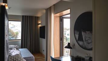 Habitación de hotel con cama, escritorio, silla, ventana grande con cortinas y un cuadro en la pared.