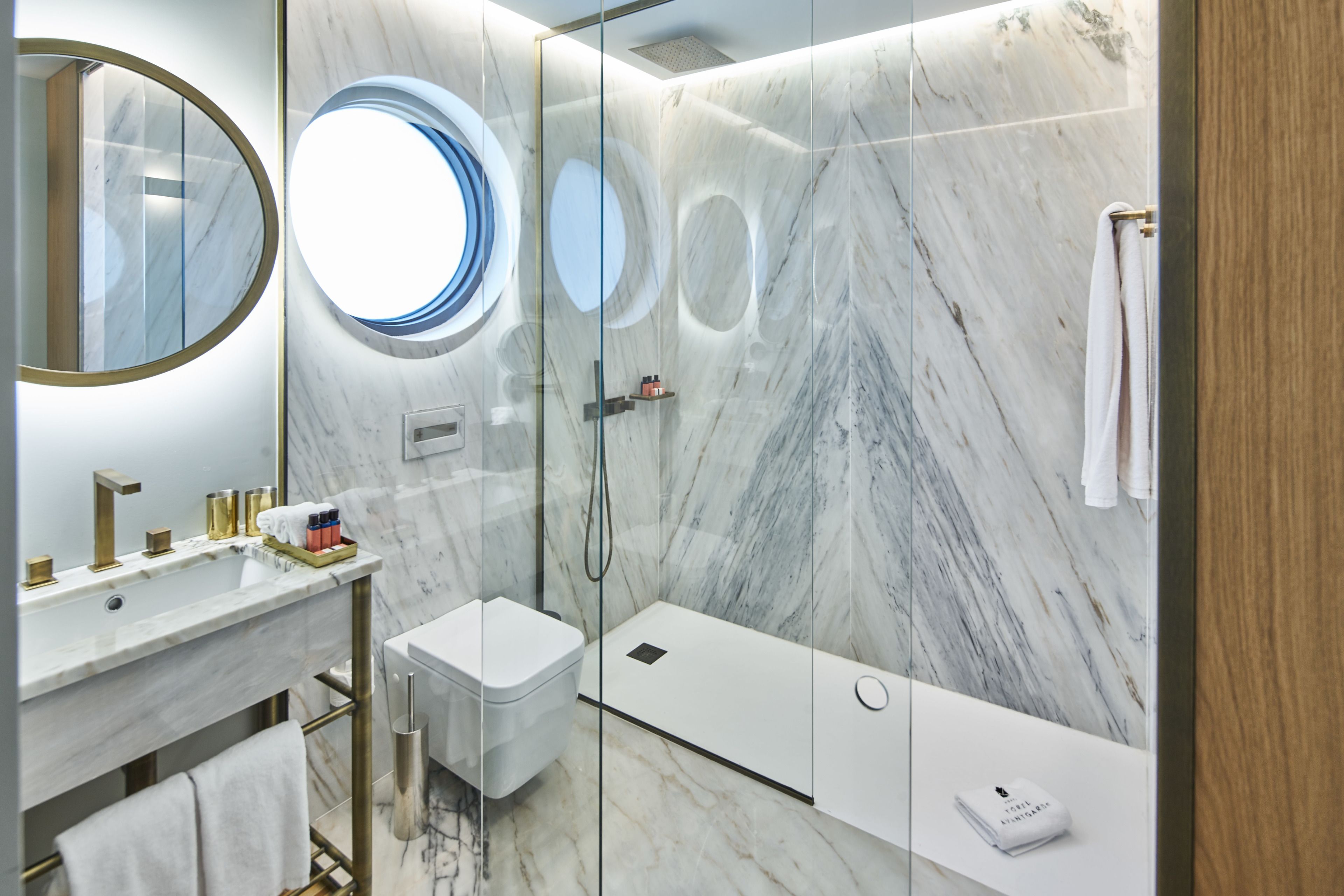A modern bathroom with a marble wall, a round mirror, a toilet, a sink, and a bathtub.