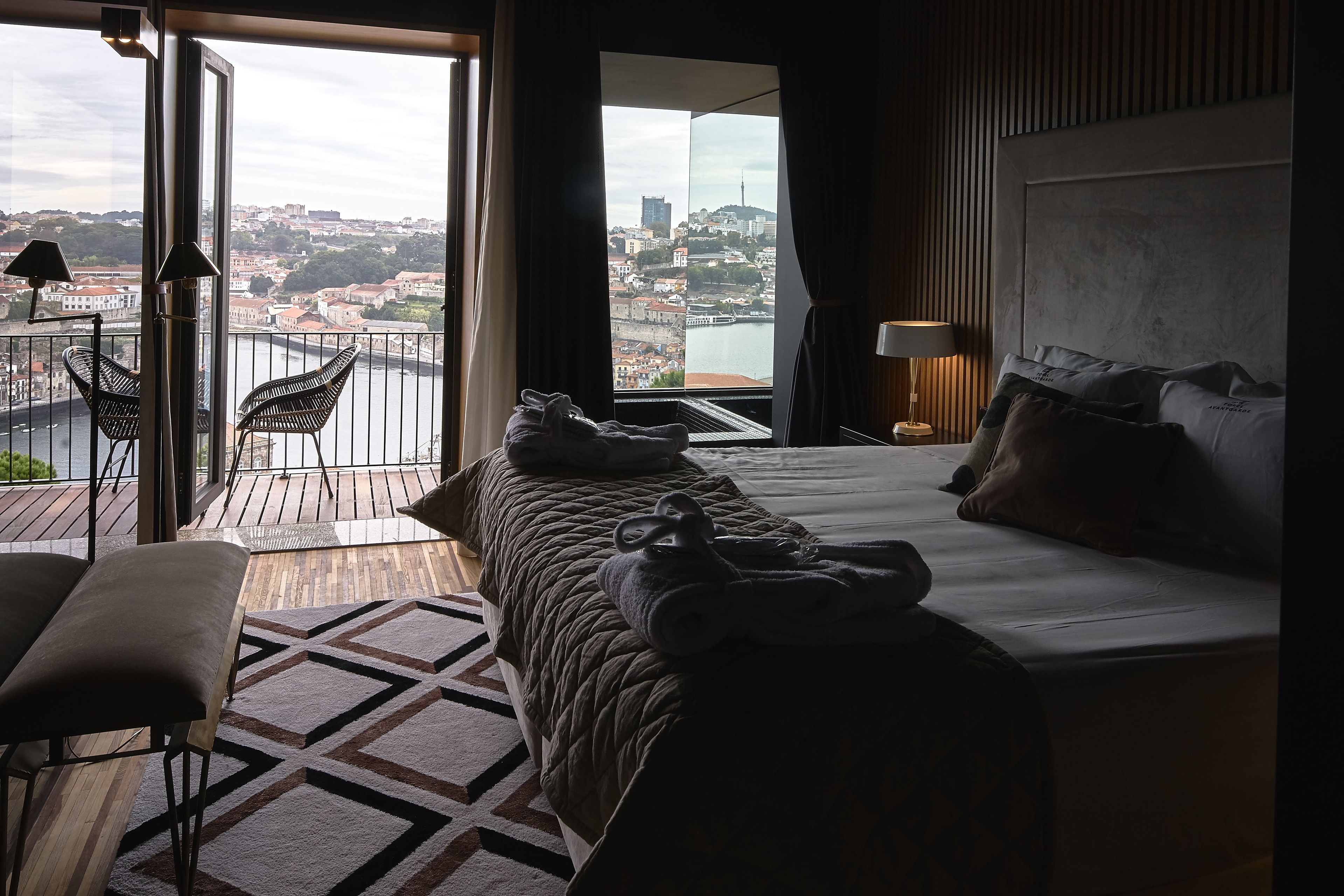 A hotel room with a large bed, a view of a cityscape, and a balcony with chairs.