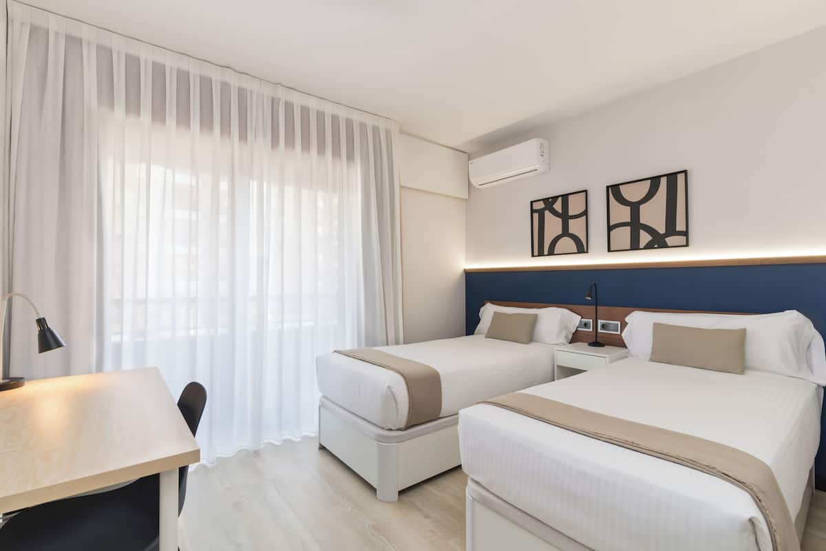 Ein Hotelzimmer mit zwei Betten, einem Schreibtisch, einem Stuhl, einem Nachttisch und einer Klimaanlage.