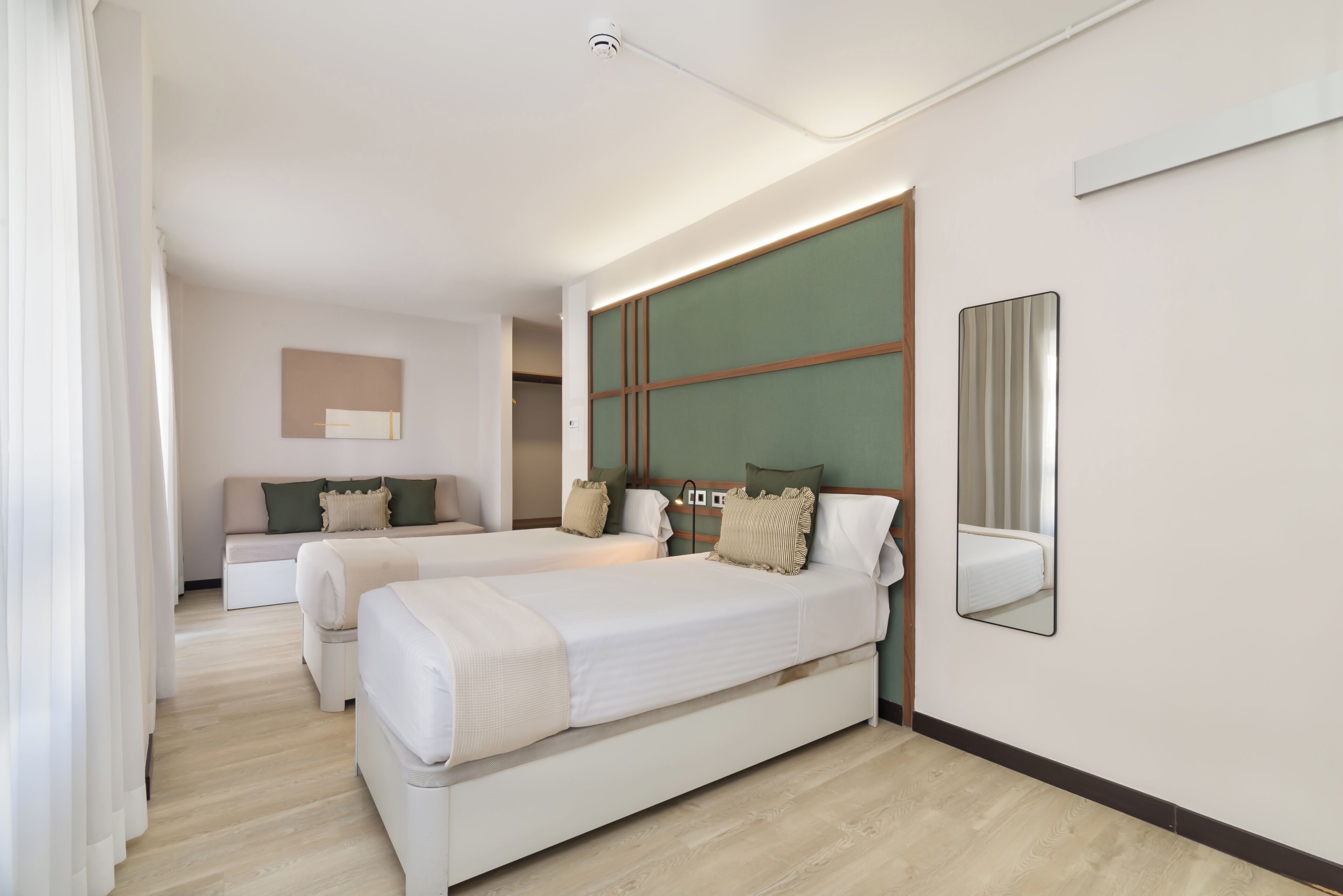 A hotel room with two single beds, a sofa bed, a large mirror, and a view of the hallway.