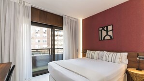 Ett hotellrum med en stor säng, en sänggavel i trä, utsikt över byggnader genom en skjutdörr och ett litet runt bord med en lampa.