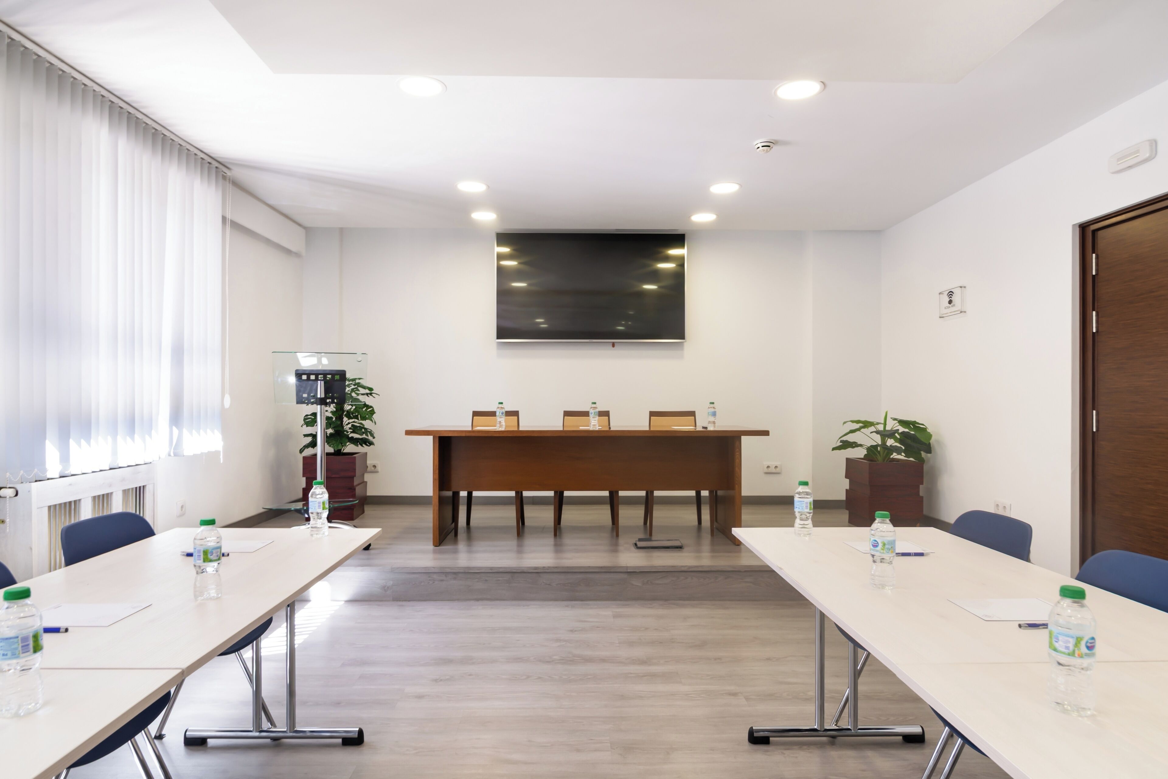 Un salón de conferencias con una larga mesa, sillas, botellas de agua, una planta y un amplio ventanal con persianas.