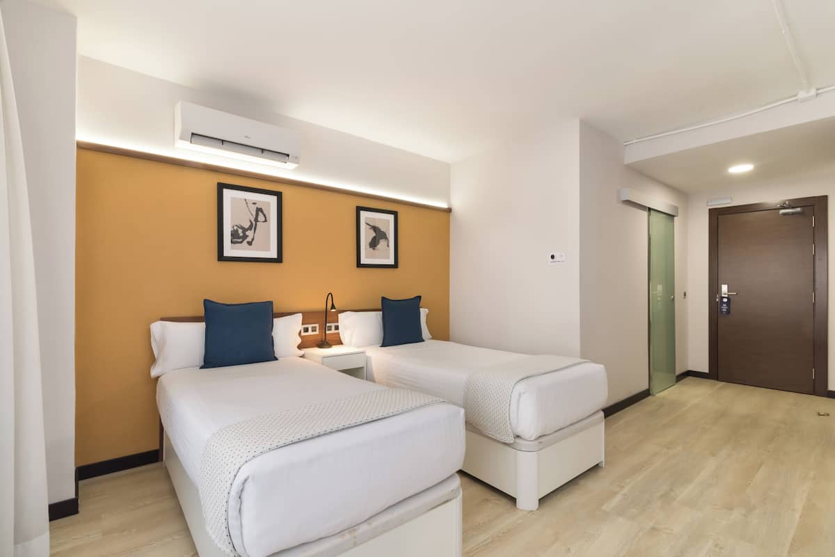Ein Hotelzimmer mit zwei Einzelbetten, einem Nachttisch, einer Wand-Klimaanlage und einer Tür mit Griff.