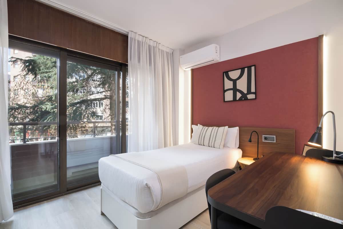 Ein Hotelzimmer mit einer großen Schiebeglasdoor zum Balkon, einem Bett mit weißer Bettwäsche, einem Holztisch, einem Nachttisch und einem modernen Wandbild.