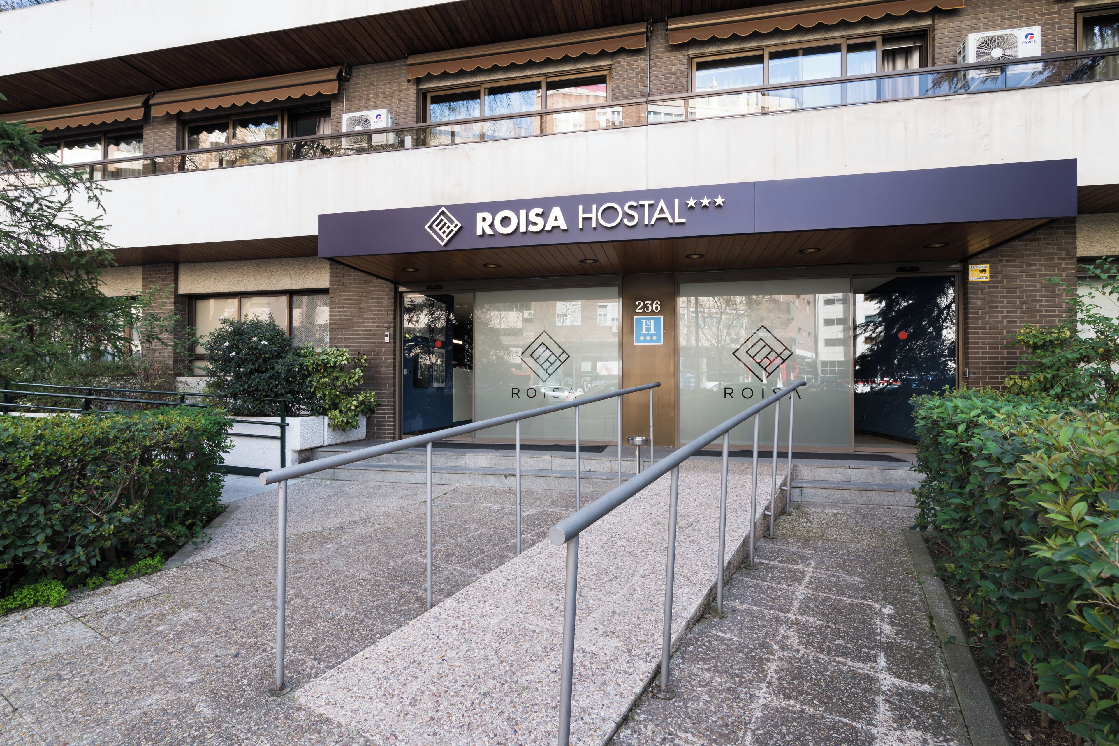 The exterior of a hotel named ROISA HOSTAL with a modern design, featuring a prominent entrance with glass doors and a metal staircase leading up to the entrance.