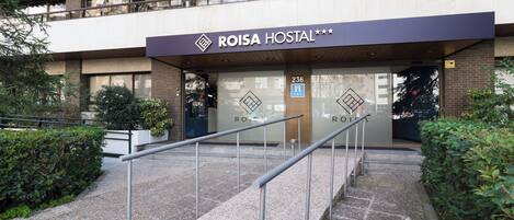 The exterior of a hotel named ROISA HOSTAL with a modern design, featuring a prominent entrance with glass doors and a metal staircase leading up to the entrance.