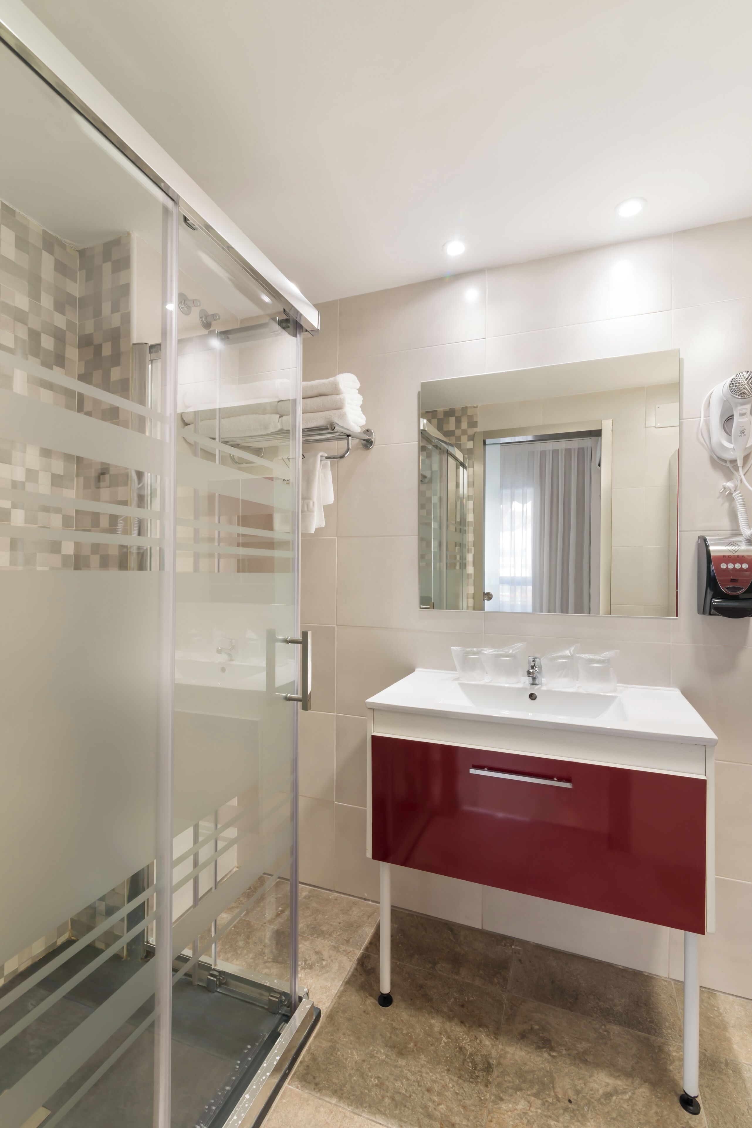 A modern bathroom with a glass shower enclosure, a white sink with a red cabinet, and a hairdryer mounted on the wall.