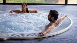 Un jacuzzi con dos personas adentro, rodeado por un ventanal grande con vistas a un bosque.