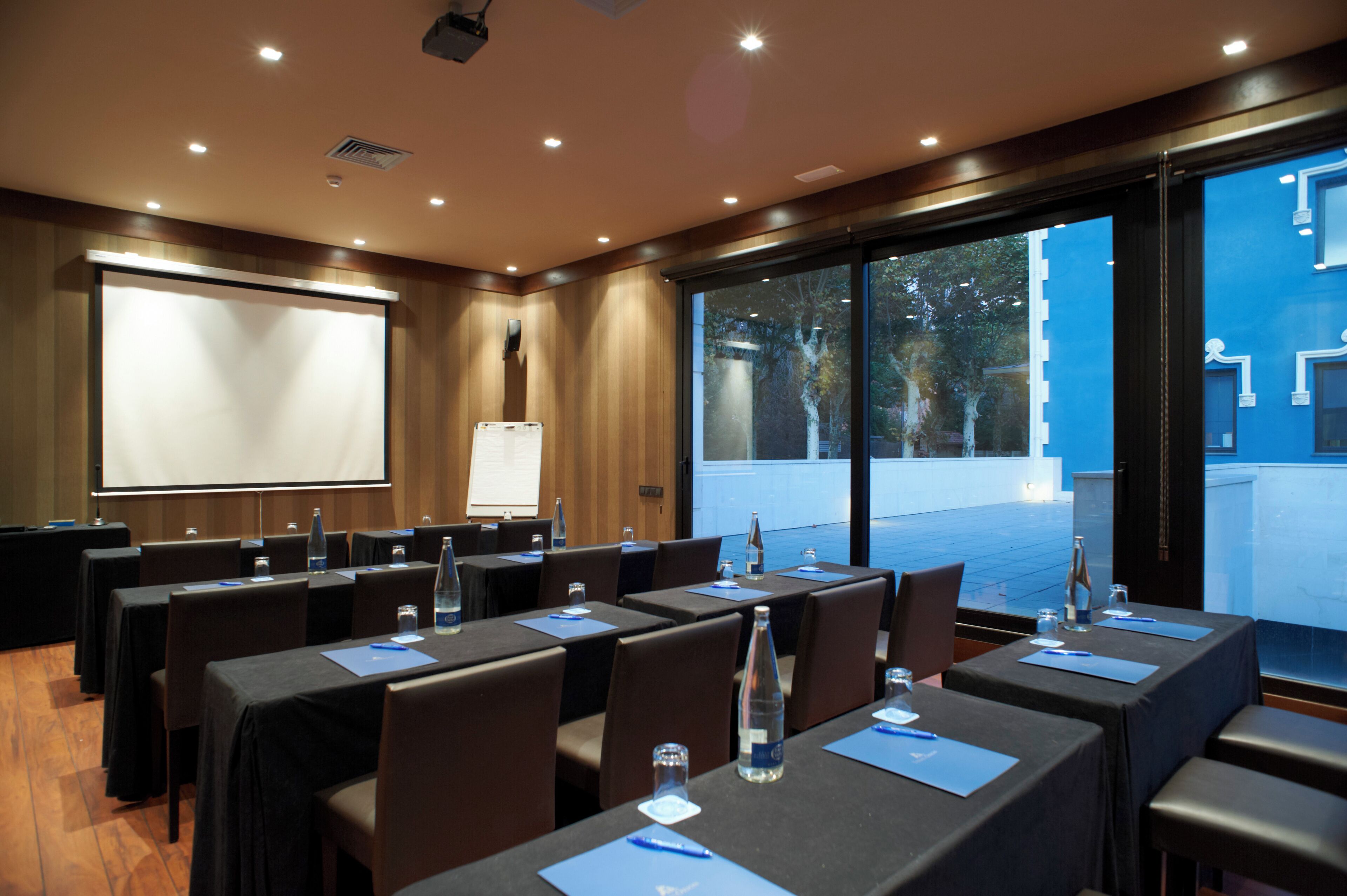 A conference room with tables, chairs, a projector screen, and a large window with a view of the outside.