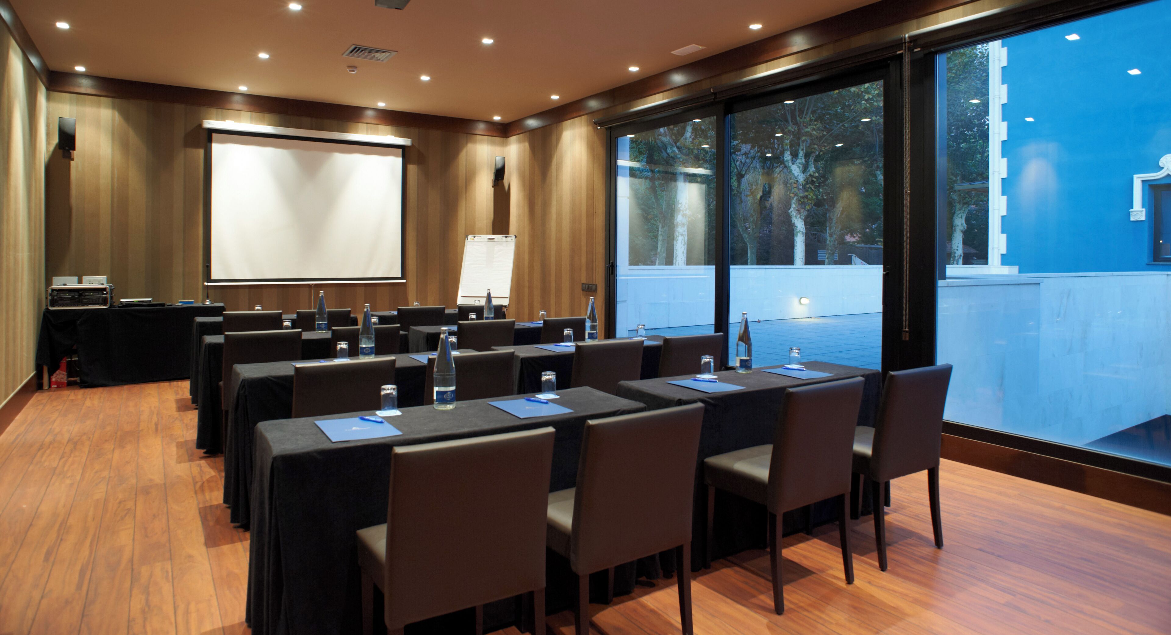 Un salón de conferencias con mesas, sillas, una pantalla de proyector y vistas al exterior.