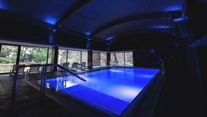 Una piscina interior con techo iluminado en azul, paredes de vidrio transparente y vistas al exterior.