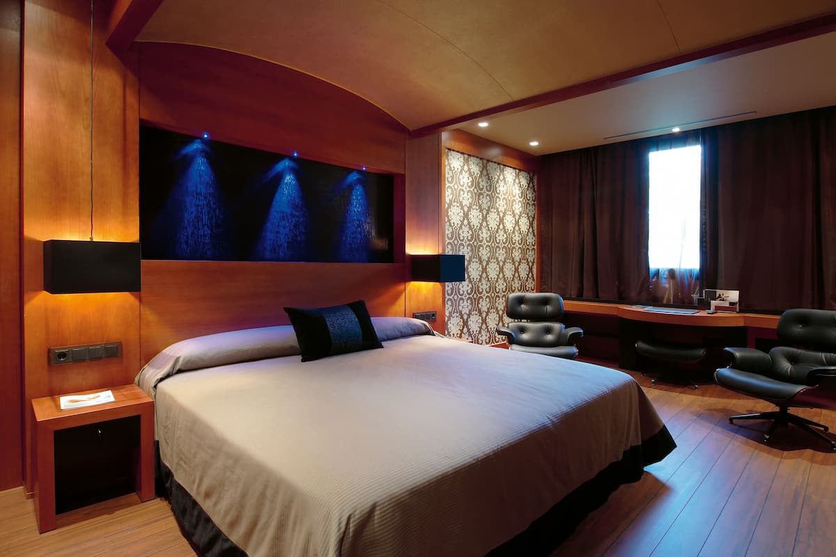 A hotel room with a large bed, a desk, two chairs, and a wall-mounted light fixture.
