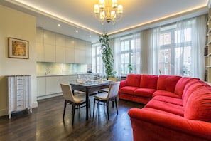 Un salon moderne avec un canapé rouge, une table à manger et des chaises. On aperçoit au fond une cuisine avec des meubles blancs et un lustre.