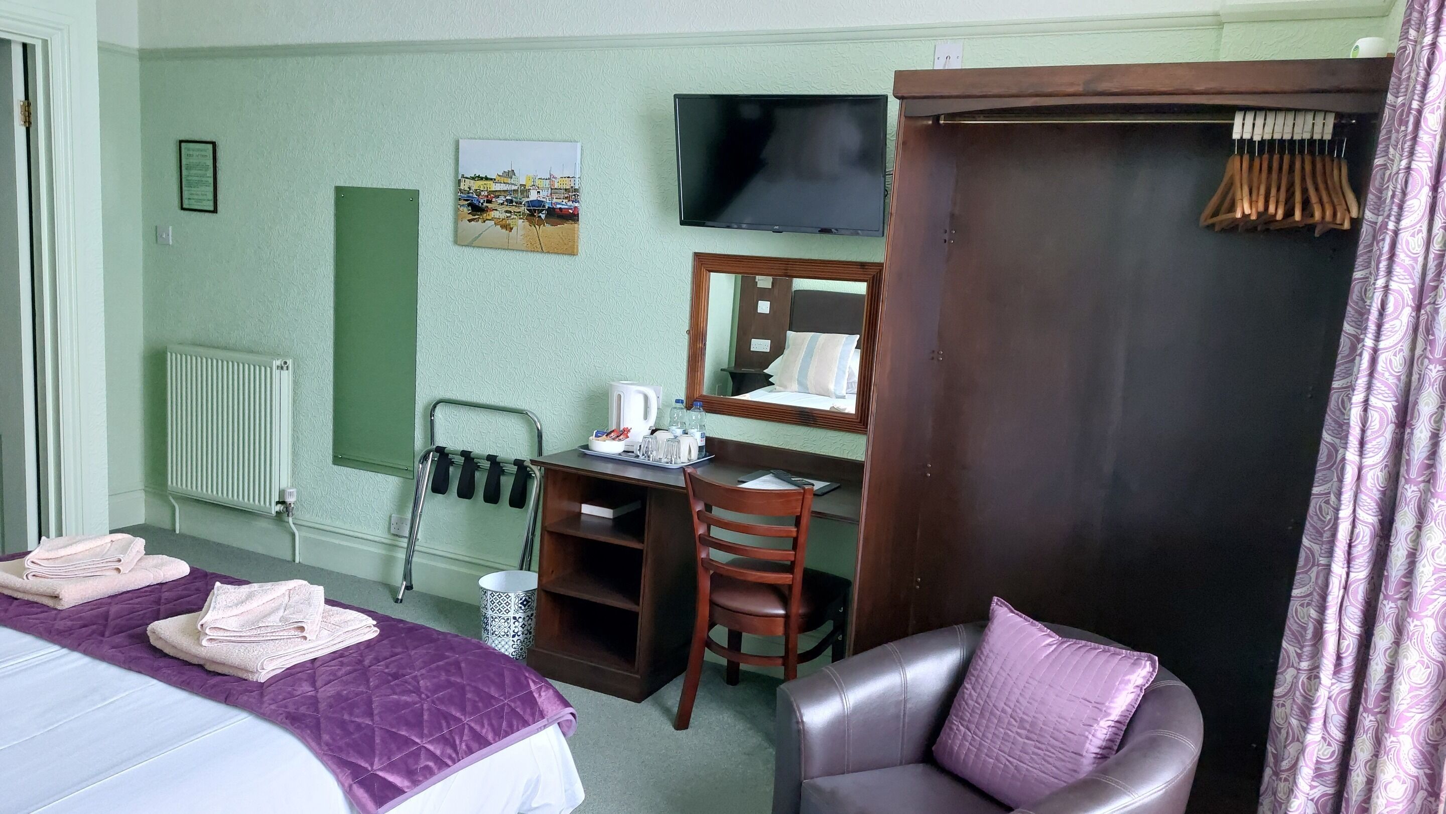 A hotel room with a bed, a desk with a chair, a television, a mirror, and a wardrobe.