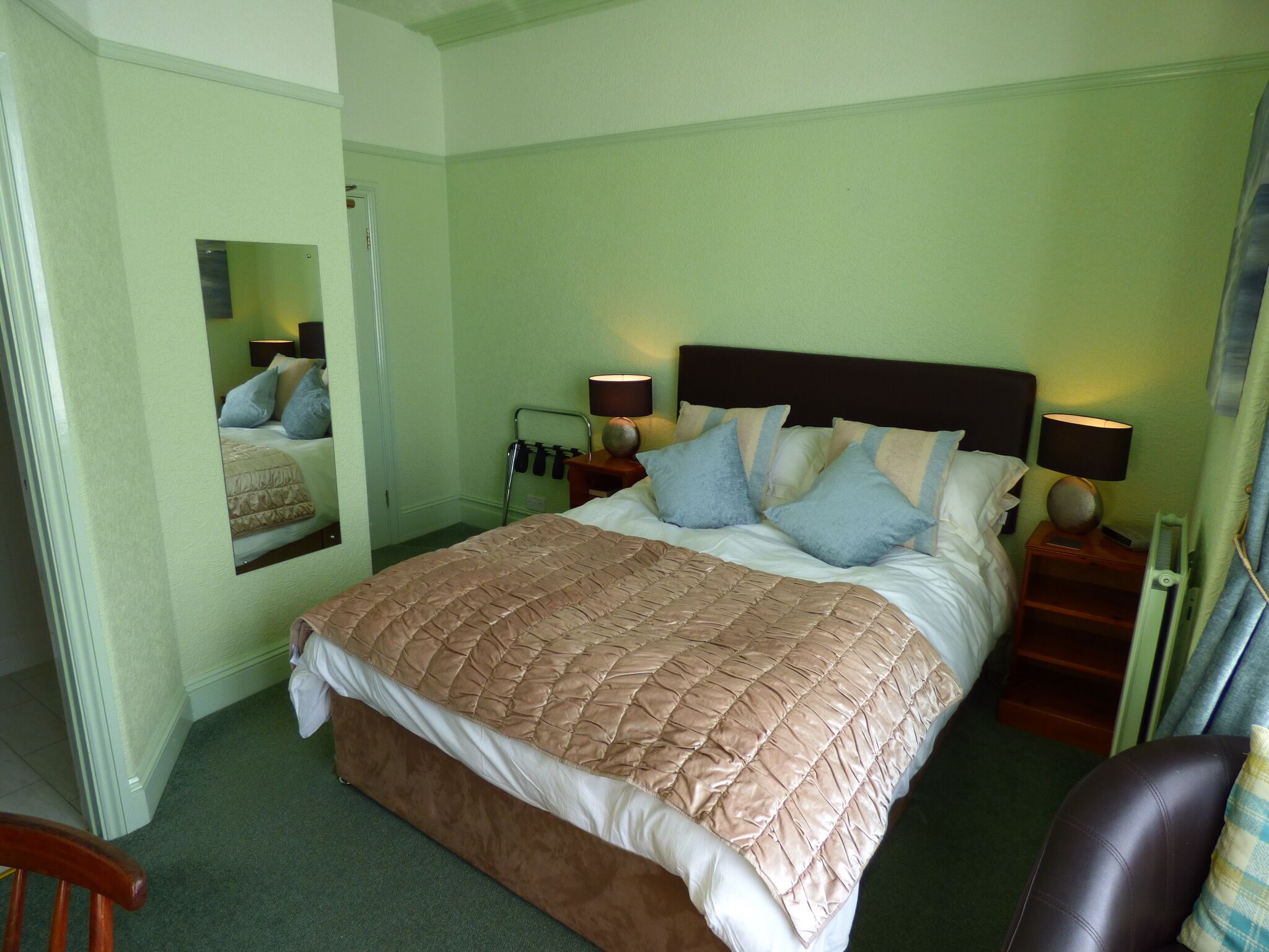 A neatly made bed with a brown quilt, two bedside tables with lamps, a mirror on the wall, and a chair.