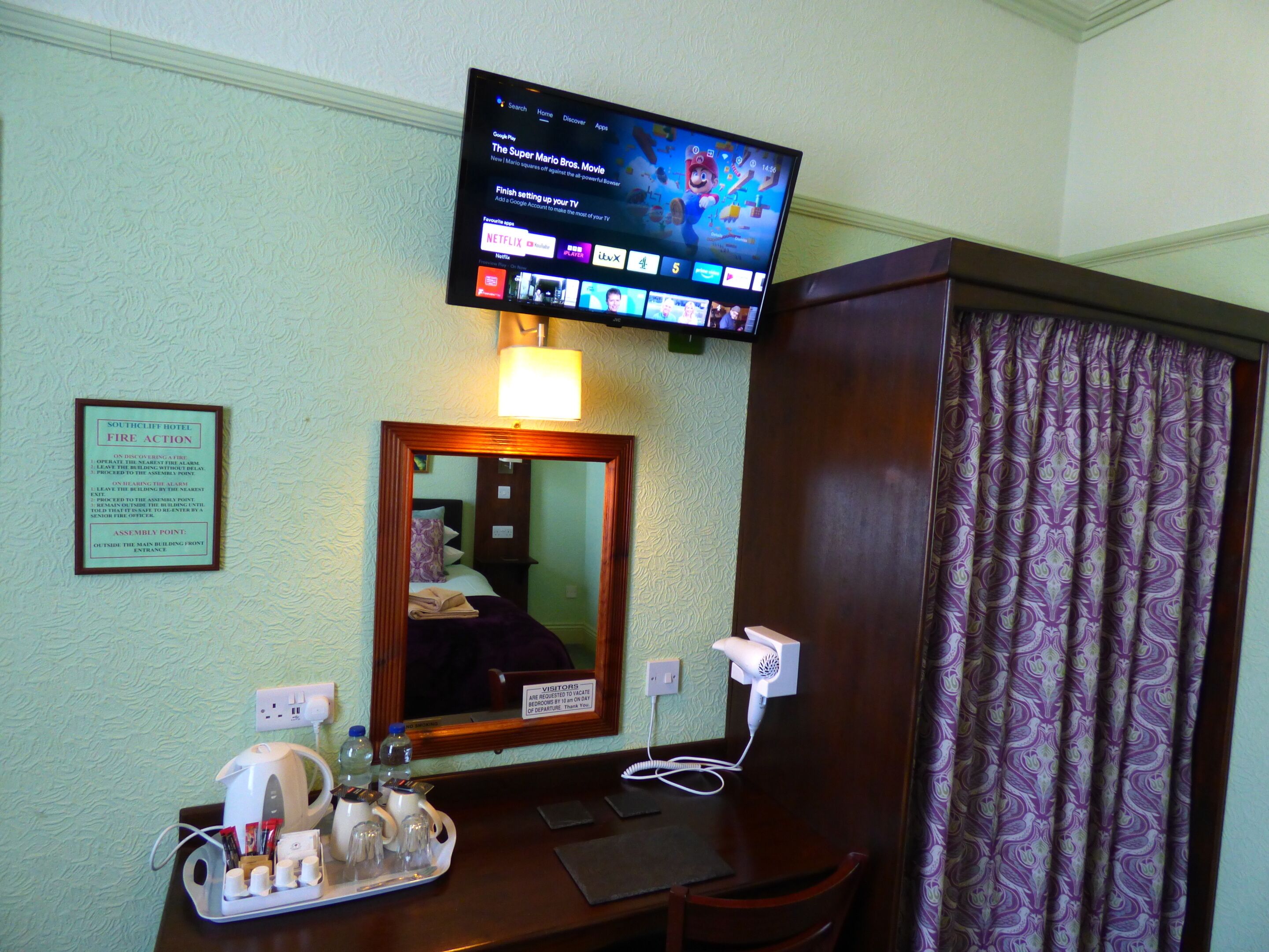 A hotel room with a television displaying a movie, a desk with a hairdryer, a mirror, and a sign indicating the assembly point for fire action.