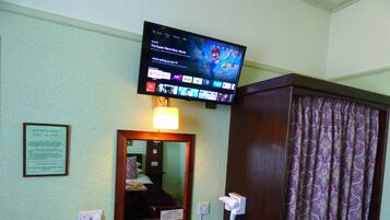 A hotel room with a television displaying a movie, a desk with a hairdryer, a mirror, and a sign indicating the assembly point for fire action.