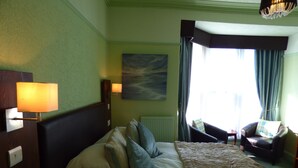 A hotel room with a large bed, two bedside lamps, a painting on the wall, a view of the outdoors through curtains, and a chair by the window.