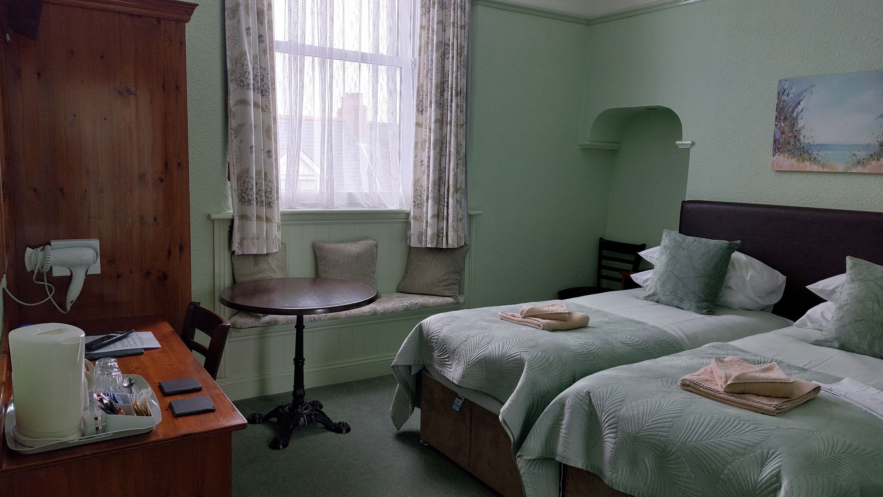 A room with two beds, a small round table, a chair, a desk with a hairdryer, and a window with curtains.