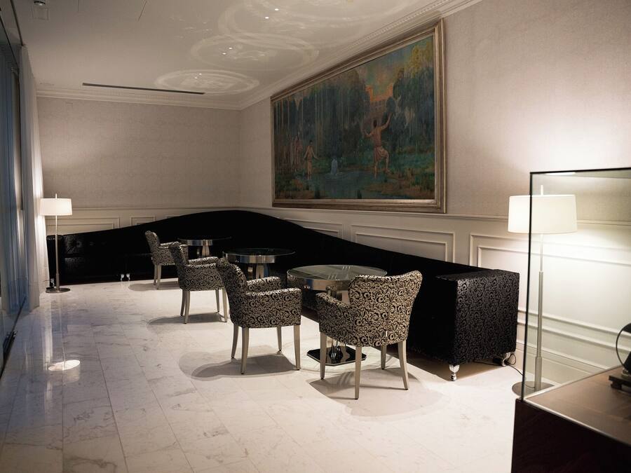 Um saguão de hotel com piso de mármore, cadeiras estampadas, uma mesa redonda, uma pintura grande e uma televisão sobre um suporte.