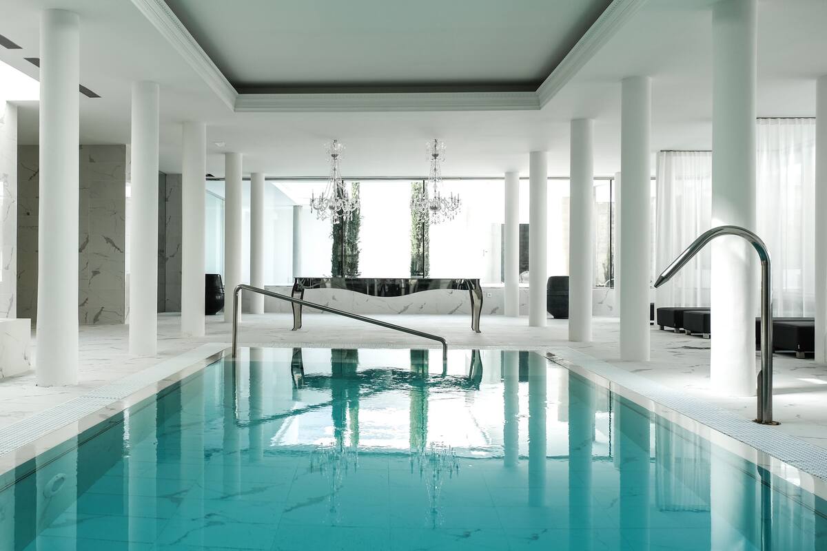 A spacious indoor swimming pool with a clear view of the ceiling and columns, a central seating area, and a modern chandelier.