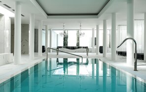 A spacious indoor swimming pool with a clear view of the ceiling and columns, a central seating area, and a modern chandelier. - Hotel Balneari Termes Orion (Santa Coloma de Farners)