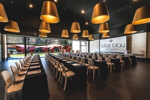 Une salle de conférence dotée de longues tables et de chaises, de grands luminaires suspendus et offrant une vue sur un jardin grâce à de larges fenêtres.