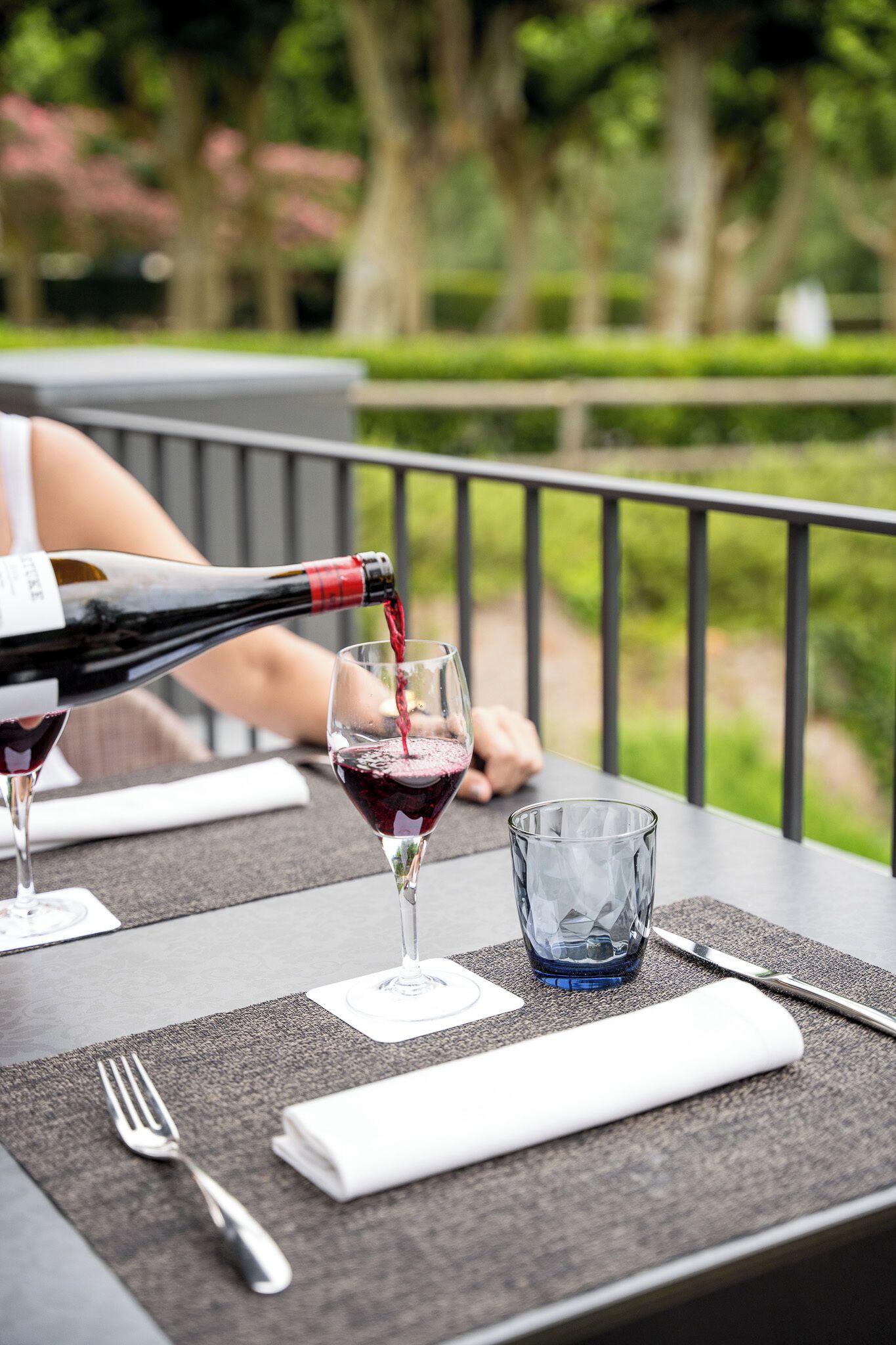Mesa al aire libre con una botella de vino tinto sirviéndose en una copa, una servilleta, un tenedor y un vaso de agua.