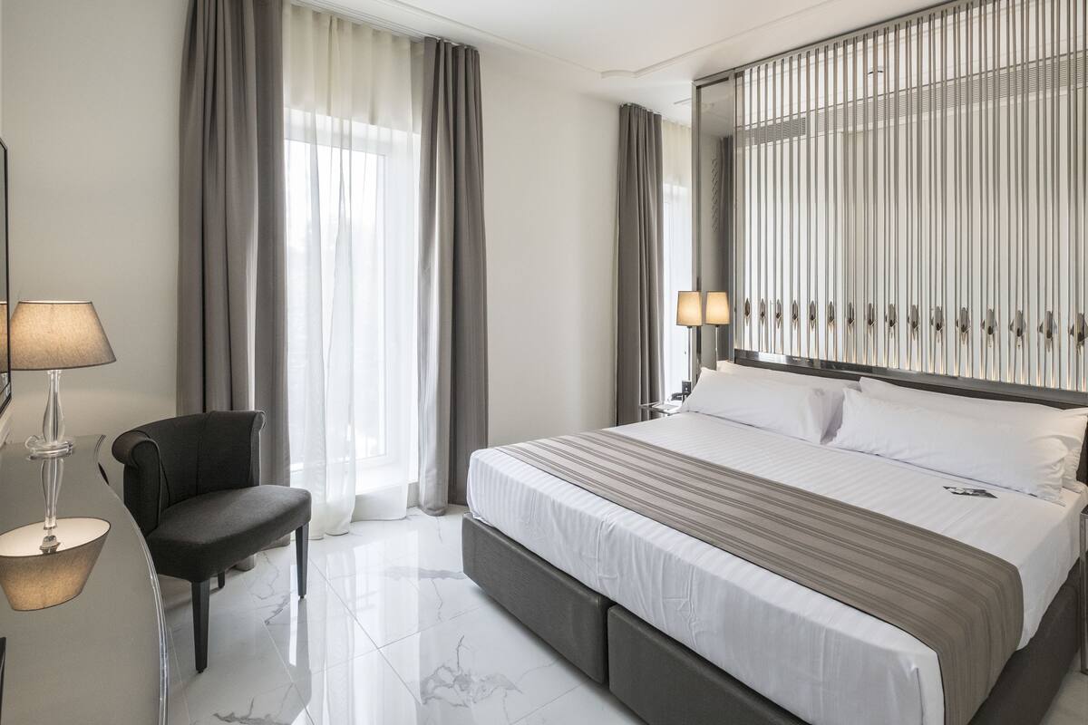 A modern hotel room with a large bed, a chair, a desk, and a mirror.